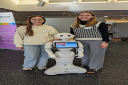 Zwei Personen posieren neben einem humanoiden Roboter. Der Roboter zeigt auf seinem Bildschirm verschiedene Auswahlmenüs.
