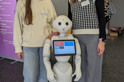 Zwei Personen posieren neben einem humanoiden Roboter. Der Roboter zeigt auf seinem Bildschirm verschiedene Auswahlmenüs.