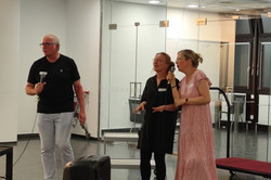 Auf dem Bild stehen drei Personen und halten Mikrofone in den Händen. Sie singen Karaoke. Eine Person trägt ein schwarzes T-Shirt und helle Hosen, während eine andere in einem schwarzen Kleid steht. Die dritte Person hat ein rosafarbenes, gemustertes Kleid an und trägt weiße Schuhe. Im Vordergrund ist ein Lautsprecher sichtbar.