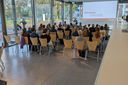 Mehrere Personen sitzen in Reihen auf Holzstühlen und hören einem Vortrag zu. Auf der Leinwand steht „Blick auf heute – Technologie und Kommunikation und Kommunikationstechnologie“.
