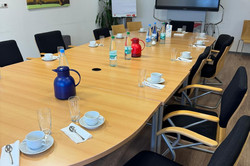 Das Bild zeigt einen Konferenzraum mit einem großen, ovalen Tisch, der für eine Besprechung vorbereitet ist. Auf dem Tisch stehen mehrere Kaffeetassen und Gläser, sowie Wasserflaschen und ein roter Kaffeekanne. Im Hintergrund befindet sich ein großer Bildschirm, der für Präsentationen genutzt werden kann. An einer Wand hängt ein Flipchart, das möglicherweise für Notizen oder Diagramme verwendet wird. Ein freundliches Kunstwerk mit einem Frosch verleiht dem Raum eine angenehme Atmosphäre. 