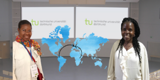 Zu sehen sind zwei Wissenschaftlerinnen welche lächelnd links und rechts vor Ihrem an eine weiße Tafel projezierten Vortrag stehen.