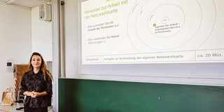 Eine junge Frau mit welligen blonden Haaren steht vor einer Tafel und schaut in die Kamera.