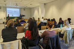 Studierende sitzen an Tischen und hören zwei Vortragenden vorne zu, die eine Präsentation über Beratung zu Studium, Beruf und Ausbildung zeigen.