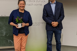 Zwei Personen stehen in einem Seminarraum vor einer Tafel und einem Roll-Up-Banner. Das Banner trägt die Aufschrift „70 Jahre rehawiss – Der Blick zurück nach vorne“ sowie „Fakultät Rehabilitationswissenschaften“. Eine Person hält eine kleine Topfpflanze in den Händen und trägt einen dunkelblauen Blazer, ein dunkles Shirt, orangefarbene Hosen und dunkelblaue Schuhe. Die andere Person ist mit einem dunkelblauen Blazer, einem hellblauen Hemd, Jeans und schwarzen Schuhen gekleidet.