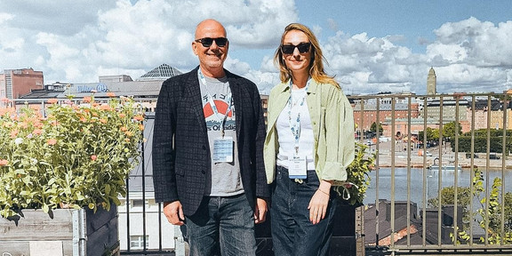Ein Mann und eine Frau mit Konferenz-Lanyards stehen lächelnd auf einer Dachterrasse, im Hintergrund die Skyline von Helsinki mit Hafen und blauem Himmel mit Wolken.