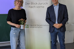 Eine blonde Frau mit Brille, schwarzem Oberteil, heller Jeans und Sneakers steht links neben einem Roll-up mit dem Schriftzug „70 Jahre reha WISS – Der Blick zurück nach vorne“. Sie hält eine weiße Tasse mit einer kleinen Pflanze. Rechts daneben steht ein Mann mit grauen Haaren und Brille, im dunkelblauen Sakko, hellblauen Hemd und Jeans. Beide Personen lächeln in die Kamera.