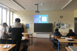 Ein Klassenzimmer mit mehreren Schüler*innen, die auf eine Präsentation schauen. Die Folie auf dem Beamer zeigt die Frage „Wie kann man sich vor Cybermobbing schützen?“. Einige Schüler*innen sitzen an Tischen, andere stehen. An der Wand hängen Sprechblasen mit positiven Botschaften und ein Plakat. Die Atmosphäre wirkt ruhig und konzentriert.