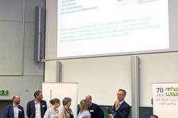 Auf dem Bild ist der Hörsaal bei der Jubiläumsfeier zu sehen, bei der ein Dankeschön für die Organisator*innen des Jubiläums und der Feier ausgesprochen wurde. Auf einer großen Leinwand im Hintergrund sind die Namen der beteiligten Gruppen und Personen aufgelistet. Vor dem Bildschirm stehen mehrere Personen auf einer kleinen Bühne, die glücklich aussehen. Einige von ihnen halten Mikrofone oder Notizen in der Hand.