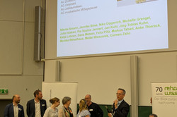 Auf dem Bild ist der Hörsaal bei der Jubiläumsfeier zu sehen, bei der ein Dankeschön für die Organisator*innen des Jubiläums und der Feier ausgesprochen wurde. Auf einer großen Leinwand im Hintergrund sind die Namen der beteiligten Gruppen und Personen aufgelistet. Vor dem Bildschirm stehen mehrere Personen auf einer kleinen Bühne, die glücklich aussehen. Einige von ihnen halten Mikrofone oder Notizen in der Hand.