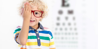 Symbolbild Augenuntersuchung Das Bild ist ein Symbolbild für eine Augenuntersuchung. Ein kleiner Junge mit lockigem blondem Haar trägt eine rote Brille und hält sich lächelnd mit einer Hand ein Auge zu. Er trägt ein bunt gestreiftes T-Shirt und steht vor einer verschwommen im Hintergrund zu erkennenden Sehtafel.