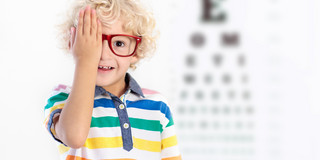 Das Bild ist ein Symbolbild für eine Augenuntersuchung. Ein kleiner Junge mit lockigem blondem Haar trägt eine rote Brille und hält sich lächelnd mit einer Hand ein Auge zu. Er trägt ein bunt gestreiftes T-Shirt und steht vor einer verschwommen im Hintergrund zu erkennenden Sehtafel.