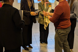 Mehrere Personen stehen in einem Raum der Technischen Universität Dortmund bei einer feierlichen Übergabe von Abschlussurkunden. Eine Person überreicht Blumen und Taschen an zwei andere, die Urkunden in den Händen halten.