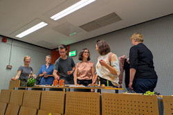 Eine Gruppe von Personen steht in einem Raum an einem Tisch, der mit verschiedenen Snacks und Getränken gedeckt ist. Auf dem Tisch liegen unter anderem Karottenstücke, Trauben und andere kleine Speisen.