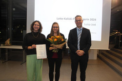 Zwei Frauen und ein Mann (rechts) schauen feierlich in die Kamera. Die Person in der Mitte hält einen Blumenstrauß in den Händen, die Person links eine Urkunde.