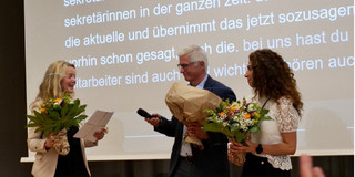 Frau Dr. Susanne Dirks, Herr Prof. Dr.-Ing. Christian Bühler und Frau Gülistan Demirbilek stehen auf einer Bühne. Sie halten jeweils einen Blumenstrauß in der Hand. Sie lächeln sich an.