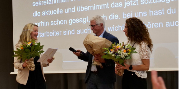 Frau Dr. Susanne Dirks, Herr Prof. Dr.-Ing. Christian Bühler und Frau Gülistan Demirbilek stehen auf einer Bühne. Sie halten jeweils einen Blumenstrauß in der Hand. Sie lächeln sich an.