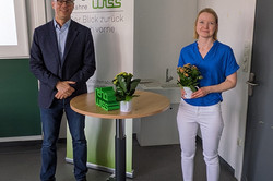 Ein Mann und eine Frau stehen nebeneinander an einem kleinen runden Tisch mit zwei Blumentöpfen. Beide lächeln in die Kamera. Der Mann trägt ein Sakko und Jeans, die Frau ein blaues T-Shirt und eine weiße Hose. Hinter ihnen steht ein Roll-up mit dem rehawiss-Logo und dem Schriftzug „70 Jahre – Der Blick zurück nach vorne“.