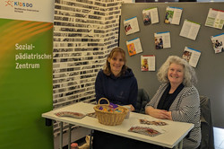 Ein Informationsstand des Klinikums Dortmund zeigt das Sozialpädiatrische Zentrum KIDS DO. Zwei Personen sitzen hinter einem Tisch mit Flyern, einer Brotschale voller Süßigkeiten sowie Infomaterialien zur Kinderförderung.