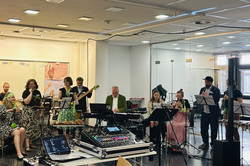 Auf dem Bild sieht man eine Gruppe von Musiker*innen, bestehend aus Mitarbeitenden der Fakultät, die in einem modernen Raum auftreten. Sie spielen verschiedene Instrumente oder singen. Einige Musiker*innen stehen, während andere sitzen. Im Vordergrund ist ein Mischpult auf einem Tisch zu sehen. Die Musiker*innen haben Notenständer vor sich und scheinen sich auf ihre Darbietung zu konzentrieren.