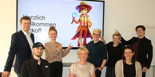 Das Projektteam KARLA bestehend aus zwei Männern und sechs Frauen präsentiert fröhlich den App-Avatar Karla: Eine Detektivin mit braunem Hut, Mantel und großer Lupe.