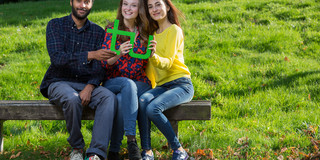 Drei Studierende sitzen auf einer Bank und halten gemeinsam ein grünes TU-Logo in den Händen.