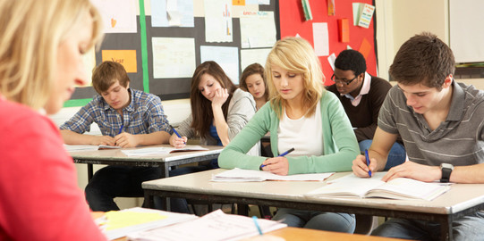 Schülerinnen und Schüler sitzen in einer Klasse und schreiben in Hefte, vor ihnen sitzt eine Lehrerin an einem Pult.