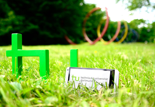 Ein Smartphone, das neben dem TU-Logo im Gras steht, im Hintergrund sind die Spektralringe zu sehen.