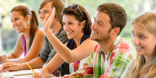 Zwei Jungen und zwei Mädchen sitzen neben einander an einem Tisch im Klassenraum und heben die Hand.