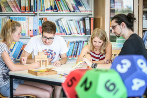 Vier Personen sitzen an einem Tisch und beschäftigen sich mit Bausteinen, die auf dem Tisch liegen. Im Vordergrund sieht man bunte vieleckige Würfel. Im Hintergund steht ein Regal voller Bücher und Ordner.