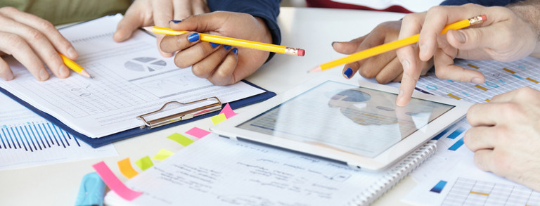 Pictured are sheets of paper and a tablet on a table. A couple of hands with pens and highlighters can be seen. Diagrams and tables can be seen on some of the paper sheets.